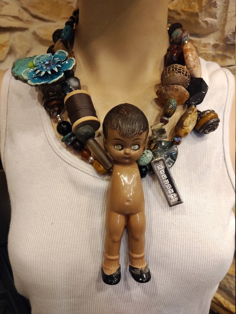May include: A multi-strand necklace with a variety of beads, including turquoise, brown, and gold. A small doll with brown hair and large eyes hangs from the center of the necklace. The doll is wearing black shoes and has a brown body.