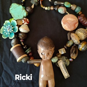 May include: A colorful beaded necklace with a variety of stones, including turquoise, carnelian, and agate. The necklace is draped over a small, brown plastic doll with large eyes. The doll is wearing a brown outfit and has the name "Ricki" written below it.