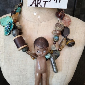 May include: A colorful necklace with a variety of beads, including turquoise, brown, and gold. A small doll with brown hair and large eyes hangs from the necklace. The necklace is on a white mannequin and a sign above reads "WE AR ART".