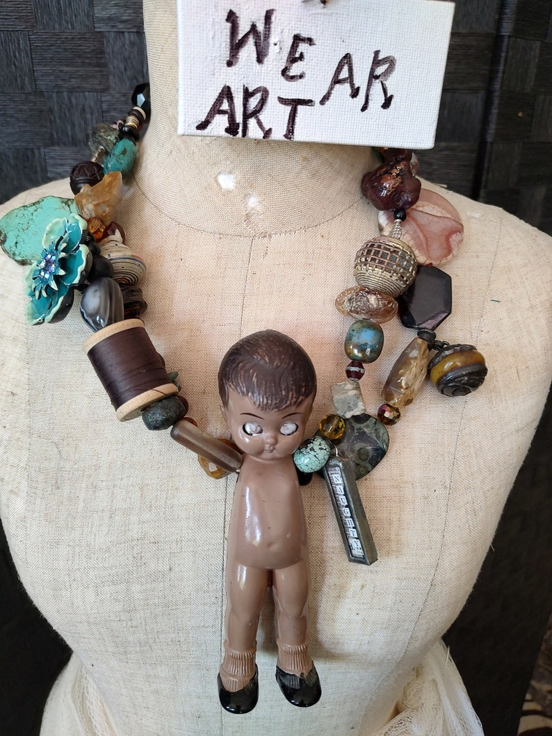 May include: A colorful necklace with a variety of beads, including turquoise, brown, and gold. A small doll with brown hair and large eyes hangs from the necklace. The necklace is on a white mannequin and a sign above reads "WE AR ART".