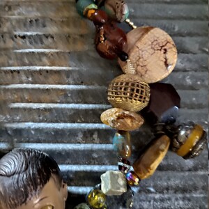 May include: A colorful beaded necklace with a variety of materials, including wood, metal, glass, and stone. The necklace features a large, round, beige stone with a cracked texture, a gold-toned wire mesh sphere, and a small, brown doll's head with closed eyes.