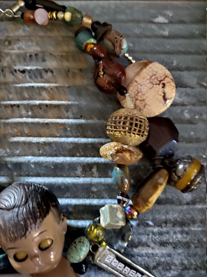 May include: A colorful beaded necklace with a variety of materials, including wood, metal, glass, and stone. The necklace features a large, round, beige stone with a cracked texture, a gold-toned wire mesh sphere, and a small, brown doll's head with closed eyes.