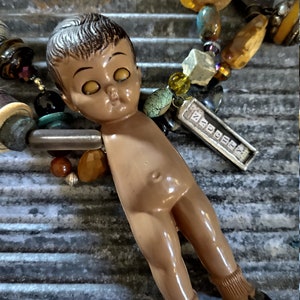 May include: A vintage doll with brown hair and large yellow eyes is part of a necklace made with various beads, stones, and charms. The necklace includes a silver charm with the word "ZODIAC" engraved on it.