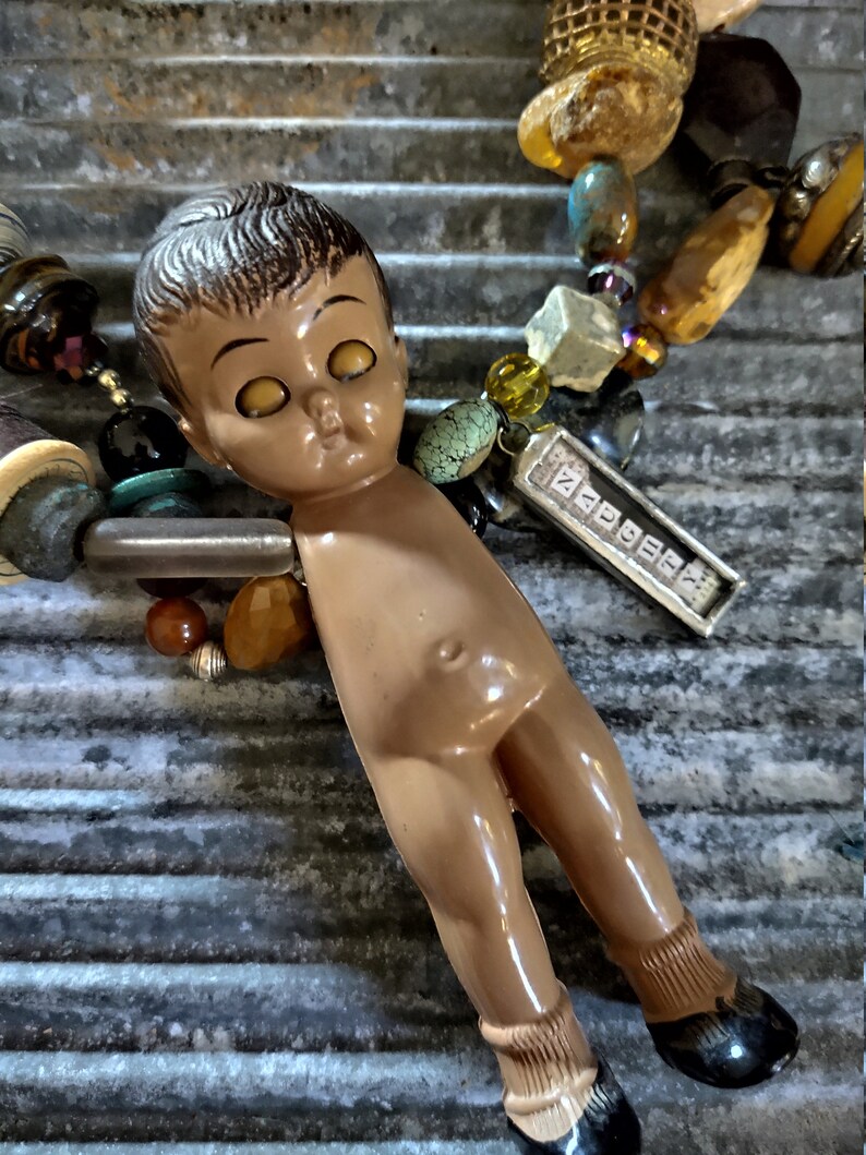 May include: A vintage doll with brown hair and large yellow eyes is part of a necklace made with various beads, stones, and charms. The necklace includes a silver charm with the word "ZODIAC" engraved on it.