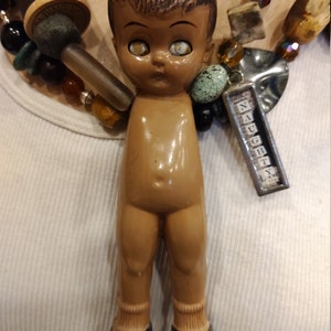May include: A vintage doll with brown hair and large eyes, wearing black shoes and socks. The doll is part of a necklace with a charm that reads "Naughty".