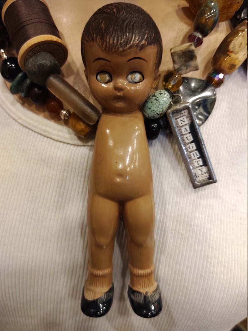 May include: A vintage doll with brown hair and large eyes, wearing black shoes and socks. The doll is part of a necklace with a charm that reads "Naughty".