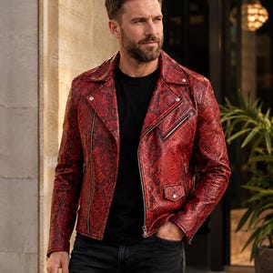 Puede incluir: Una chaqueta de motorista de cuero con estampado de piel de serpiente en rojo y negro, con solapas, cremalleras metálicas plateadas y botones a presión. La chaqueta se lleva sobre una camiseta negra lisa y unos vaqueros gris oscuro.