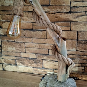 Puede incluir: Una lámpara de mesa con una base de piedra gris grande que sostiene una pieza vertical de madera flotante clara. Una cuerda gruesa de color canela está envuelta alrededor de la madera, extendiéndose desde la parte superior para sostener una bombilla ámbar.
