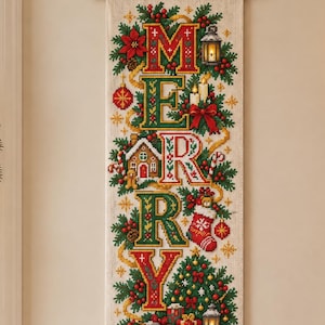 May include: A vertical wall hanging featuring cross-stitch embroidery on a light fabric background. The design displays the word MERRY in large, decorative letters surrounded by Christmas motifs including holly, a gingerbread house, a stocking, and gifts.