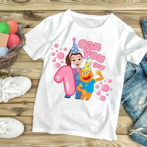 Può includere: Una t-shirt bianca con la grafica di una bambina dei cartoni animati e un mostro arancione che indossano cappellini da festa. Il design include un grande numero uno rosa, bolle rosa e il testo Can you say in corsivo rosa sul davanti.