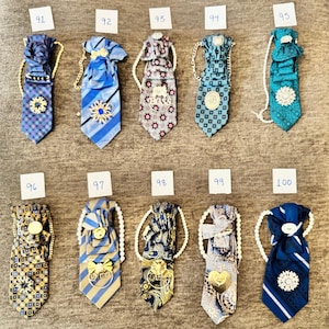 May include: A collection of ten decorative neckties labeled 91 through 100, each featuring unique fabric patterns, pleated knots, and embellishments such as metallic brooches, gold-tone charms, and white beaded or gold chain neck straps.