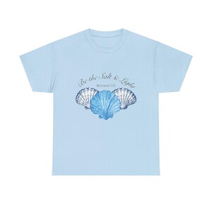 May include: A light blue short-sleeve t-shirt featuring a graphic of three blue seashells arranged in a row. Above the shells, the text reads Be the Salt & Light and Matthew 5:13 in a dark, elegant, cursive and serif font combination.