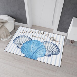 Könnte beinhalten: Eine rechteckige Fußmatte mit hellblau-weißem vertikalem Streifenmuster. Drei muschelförmige Motive im Aquarellstil in Blautönen sind unten zentriert. Der Text "Be the Salt & Light" und "MATTHEW 5:13" ist in Braun aufgedruckt.