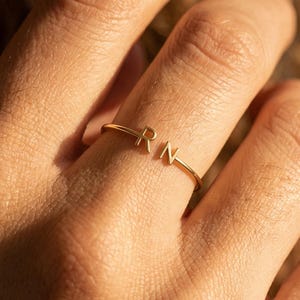 Peut inclure: Une bague ouverte fine de couleur dorée présentant deux lettres majuscules, R et N, positionnées aux extrémités de l'anneau. La bague possède une finition lisse et polie, avec les lettres se faisant face au niveau de l'ouverture.