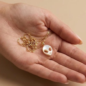 May include: A gold chain necklace featuring a pendant carved from iridescent white shell into the shape of an alien head with two large oval eye cutouts and a small mouth opening, suspended from a delicate gold-toned metal link chain.