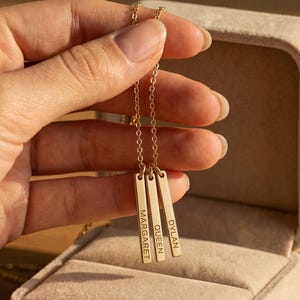 May include: A gold-toned necklace featuring three vertical bar pendants suspended from a delicate chain. Each rectangular bar is engraved with a name: MARGARET, QUEEN, and DYLAN. The jewelry is presented in a beige velvet-lined display box.