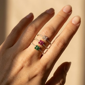 May include: Three thin gold rings, each featuring a small four-petaled flower-shaped cluster of gemstones. The top ring has clear stones, the middle ring has pink stones, and the bottom ring has green stones, all set on delicate bands.