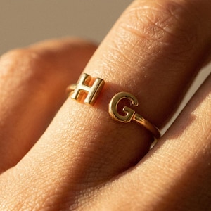 Op de afbeelding: Een goudkleurige open ring met twee hoofdletters, H en G, geplaatst aan de uiteinden van de band. De ring heeft een gepolijste metalen afwerking en een eenvoudig, dun bandontwerp dat gedeeltelijk om de vinger wikkelt en de letters toont.