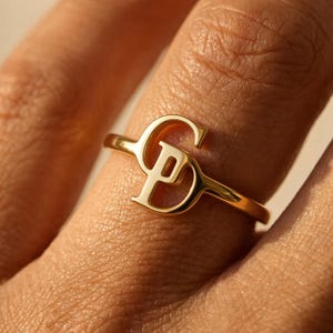 May include: A gold ring featuring a polished finish with two interlocking letters, S and P, forming the central design element. The band is thin and circular, showcasing a smooth metallic surface that reflects light across the letter shapes.