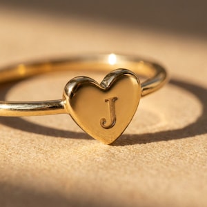 Op de afbeelding: Een goudkleurige ring met een glad, gepolijst hartvormig middendeel. In het midden van het hart is de hoofdletter J gegraveerd. De ringband is dun en rond, met een consistente metalen afwerking over het gehele ontwerp.