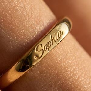 Peut inclure: Une bague en or poli portant le prénom Sophia gravé dans une police d'écriture cursive. La surface métallique est lisse et réfléchissante, mettant en valeur les lettres détaillées gravées directement sur l'anneau de la bague portée au doigt.