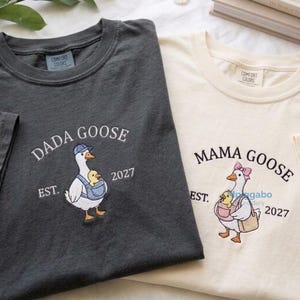 May include: A charcoal grey t-shirt and a cream t-shirt, both featuring embroidered designs. The grey shirt displays a goose in a blue cap and carrier with the text DADA GOOSE EST. 2027. The cream shirt shows a goose in a pink bow with the text MAMA GOOSE EST. 2027.