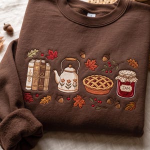 Peut inclure: Un sweat-shirt marron foncé à col rond orné de broderies représentant une pile de trois livres liés par une ficelle, une théière à motifs floraux, une tarte à croisillons et un pot étiqueté JAM, entourés de feuilles d'automne, de glands et de baies.