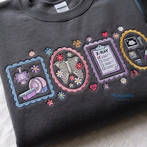 以下が含まれることがあります： 医療機器と解剖図のカラフルな刺繍が施されたダークグレーのトレーナー。CTスキャナー、肋骨、レントゲン検査報告書、超音波診断装置のデザインが、スカラップ状の縁取りと小さな花のアクセントで囲まれています。