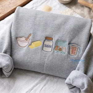 May include: A heather gray sweatshirt featuring embroidered baking ingredients including a bowl with a spoon, a stick of butter, a jar labeled SUGAR, a carton of eggs, and a measuring cup. The embroidery is centered on the front of the garment.