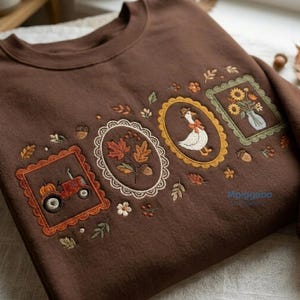 Könnte beinhalten: Ein dunkelbraunes Sweatshirt mit Rundhalsausschnitt und aufwendigen Stickereien von drei gerahmten Motiven: ein roter Traktor mit Kürbis, Herbstblätter, eine Gans mit Hut sowie eine Vase mit Sonnenblumen. Der Text Mpiggabo Embroidery ist sichtbar.