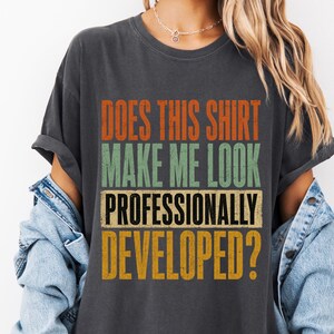 May include: A charcoal gray short-sleeve t-shirt featuring the text "DOES THIS SHIRT MAKE ME LOOK PROFESSIONALLY DEVELOPED?" in stacked, distressed-style orange, green, and yellow lettering. The shirt is worn with a light blue denim jacket.