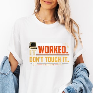 May include: A white short-sleeve t-shirt featuring a graphic print of a vintage computer icon and the text IT WORKED. DON'T TOUCH IT. in orange and black, accompanied by binary code and horizontal lines, worn with a blue denim jacket.