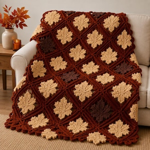 May include: A crochet blanket featuring a repeating pattern of maple leaf motifs in shades of cream, dark brown, and rust orange. The leaves are arranged in a diamond grid structure with a scalloped border edge, creating a textured textile design.