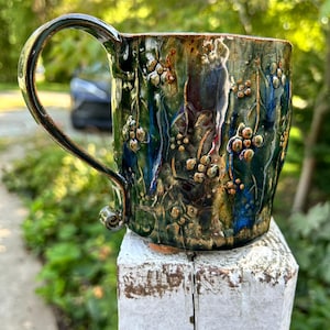 May include: A ceramic mug with a glossy, multi-colored glaze in shades of deep green, blue, and bronze. The surface features a raised, textured floral pattern with small berries and leaves. It has a large, curved handle with a curled base.