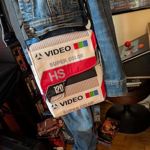 May include: A beige and red crossbody bag designed to resemble a VHS tape. The bag features the words "VIDEO SUPER COLOR HS 120" printed on the front. The bag is worn over a denim jacket and jeans.