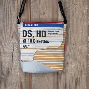 May include: A rectangular crossbody bag made from a repurposed 5 1/4" floppy disk box. The bag features a white and gold striped pattern with text that reads "FORMATTED DS, HD 10 Diskettes Double Sided, High Density". It has a black adjustable strap.