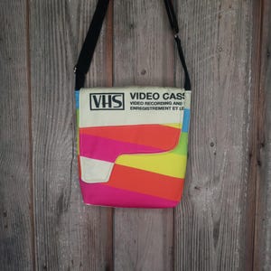 May include: A colorful messenger bag with a rainbow striped design. The bag features a flap with the text "VHS VIDEO CASSETTE VIDEO RECORDING AND ENREGISTREMENT ET LE". The bag has a black shoulder strap and a white flap.