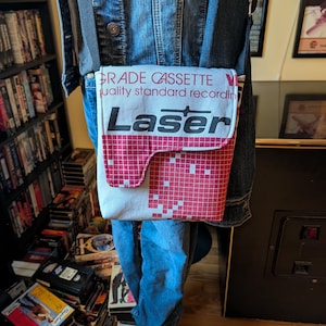May include: A crossbody bag crafted from a repurposed cassette tape case featuring a white and red grid pattern. The flap displays the text GRADE CASSETTE, quality standard recording, and the word Laser in large black letters on a white background.