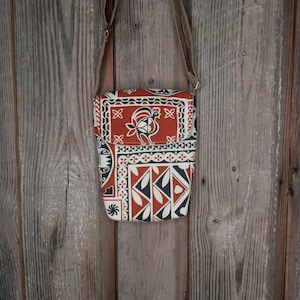 May include: A rectangular crossbody bag with a flap closure, featuring a repeating pattern in red, white, and navy blue. The bag has a brown adjustable strap and is hanging against a wooden background.
