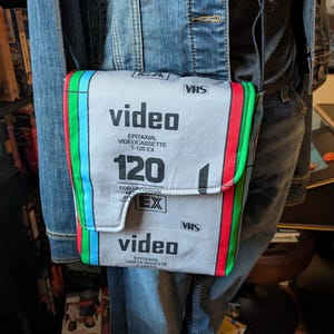 May include: A gray crossbody bag with a VHS tape design. The bag features the word "video" in large white letters, along with the text "EPITAXIAL VIDEOCASSETTE T-120 EX". The edges are trimmed with green, red, and blue stripes. It has a black strap.