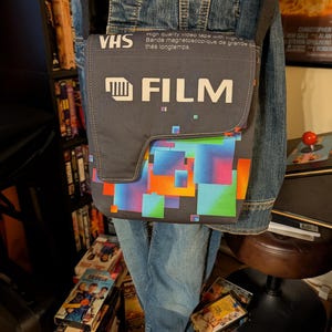 May include: Gray messenger bag with a VHS tape design. The bag features the word "FILM" in white, along with colorful geometric shapes. The bag has a black strap and is worn over a denim jacket.
