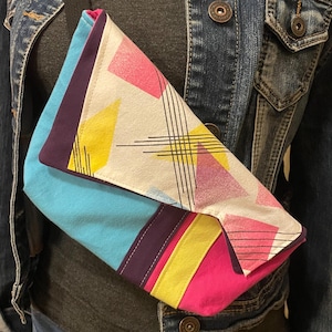 May include: A colorful crossbody bag with a geometric design. The bag features a light blue flap, a white panel with pink and yellow shapes, and a black strap. The bag is worn over a gray shirt and a denim jacket.