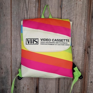 May include: A colorful backpack with a retro VHS cassette design. The bag features a rainbow striped pattern and the text "VHS VIDEO CASSETTE VIDEO RECORDING AND PLAYBACK". It has green straps and a handle, and a flap closure.