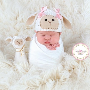 May include: A newborn baby wrapped in a white blanket, wearing a white lamb hat with pink bows and tan ears. A stuffed lamb toy with a cream bow sits nearby. The baby is lying on a fluffy white surface.