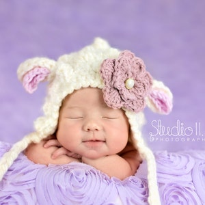 May include: A sleeping baby wearing a white crocheted lamb hat with a pink flower on top, lying on a purple textured background. The hat has pink ears and a long tail.