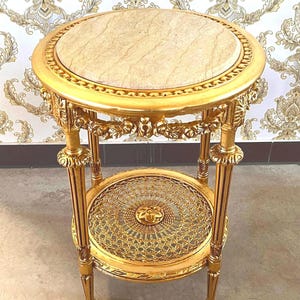 May include: A round side table with a gold-finished frame and a beige marble top. The table features four fluted legs, ornate floral carvings along the apron, and a lower circular shelf with a woven metal lattice pattern and a central floral motif.