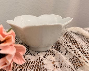 Puede incluir: Un pequeño cuenco de porcelana blanca con un borde festoneado en forma de pétalos. El cuenco presenta un acabado liso y brillante con una base circular, diseñado con una forma delicada inspirada en una flor que se curva hacia afuera.