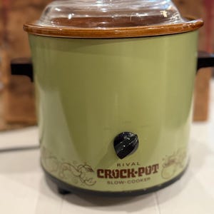 Puede incluir: Una olla de cocción lenta Rival Crock-Pot de color verde claro con borde marrón, tapa de cristal transparente y asas laterales negras. El frente presenta un mando de control negro, el texto Rival Crock-Pot Slow Cooker y un gráfico de verduras.