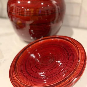 May include: A glossy, deep red lidded jar featuring a prominent circular wood grain pattern throughout its surface. The round lid is detached and positioned in the foreground, revealing its concentric ring texture, while the jar sits behind it.