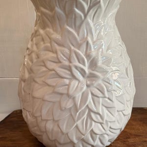 May include: A white ceramic vase with a glossy finish, featuring a textured surface covered in overlapping leaf-like or petal-shaped reliefs. The vase has a wide, scalloped rim and a rounded, bulbous body that tapers towards the base.